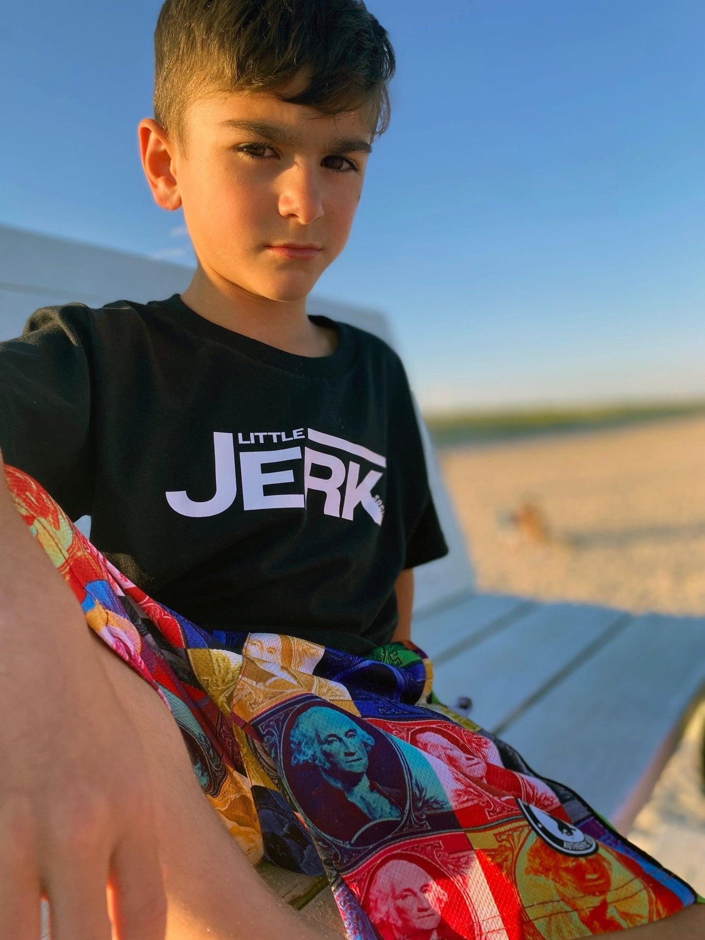 A young boy sitting on a bench wearing a black t-shirt and colorful patterned shorts.