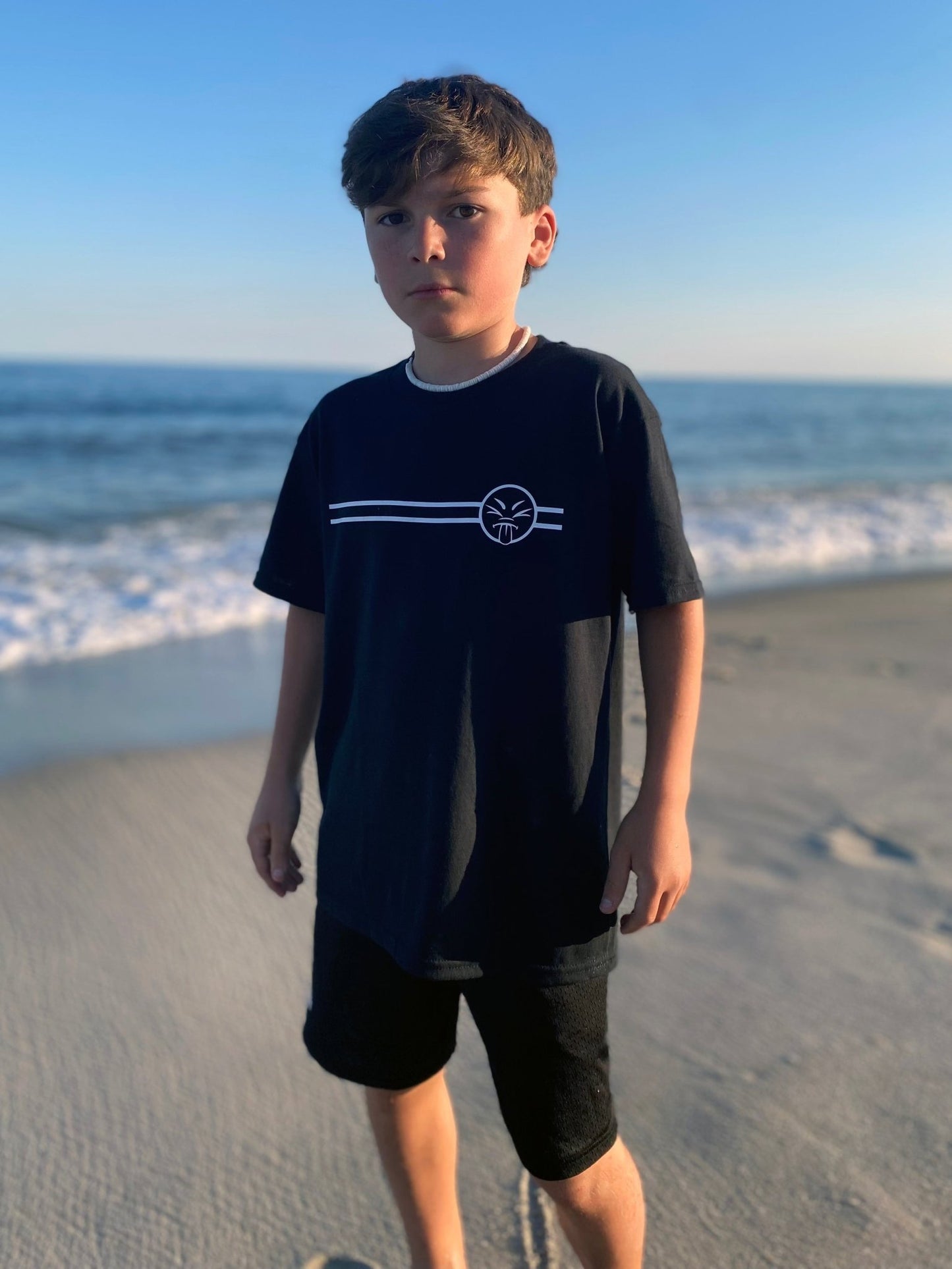A young boy standing on the beach, wearing a black t-shirt and shorts, with the sea in the background.