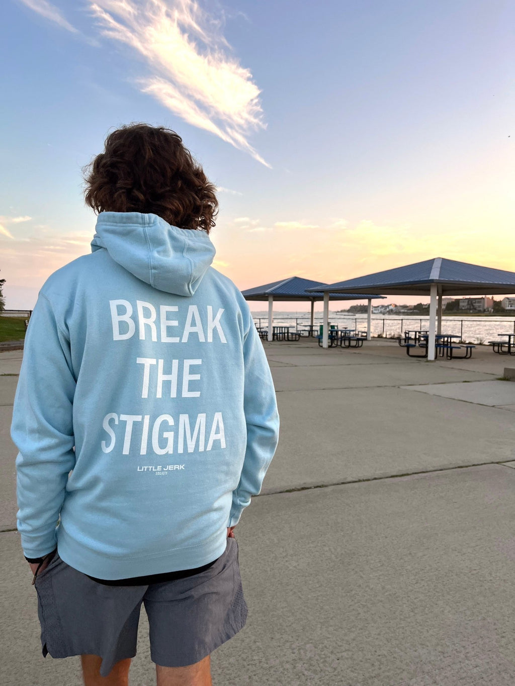 Person wearing a blue hoodie with the text 'BREAK THE STIGMA' on the back, standing outdoors by a waterfront.