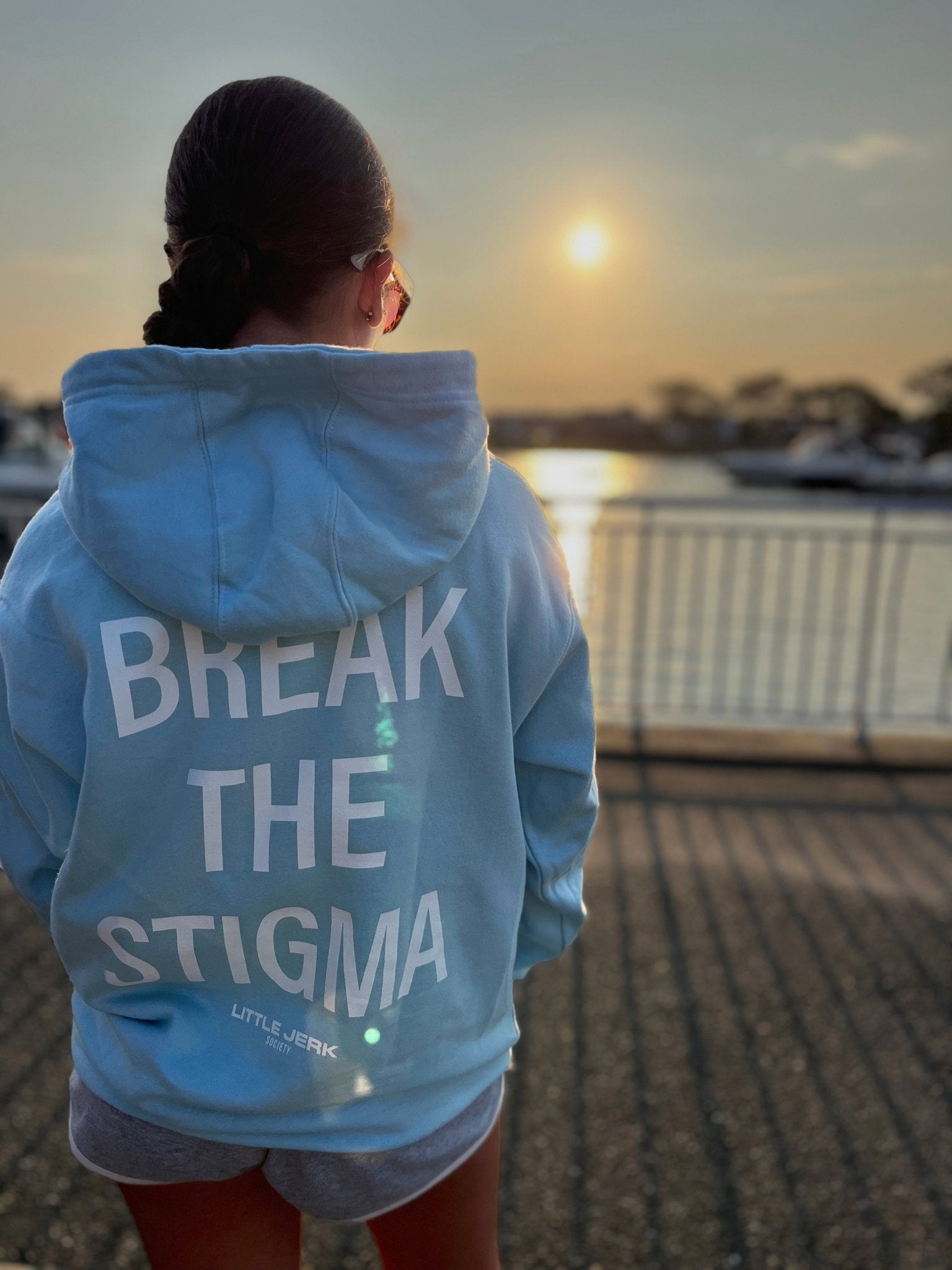 Person wearing a light blue hoodie with 'BREAK THE STIGMA' printed on the back, standing by a waterfront at sunset.