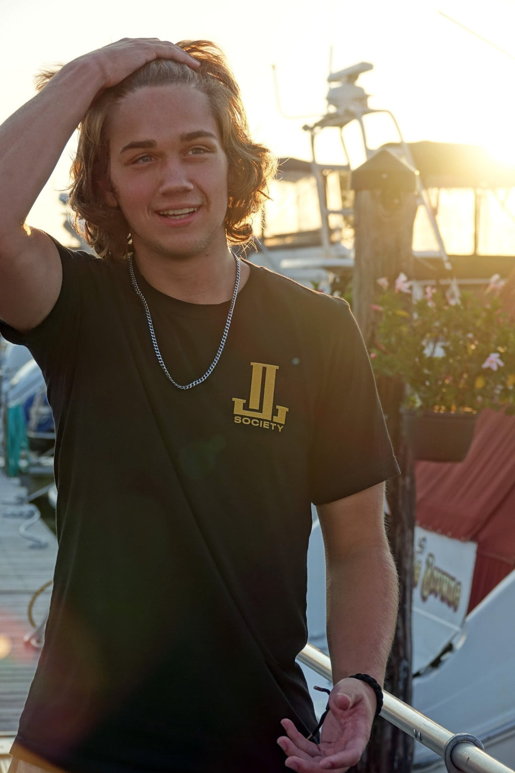 Young man with wavy hair wearing a black t-shirt, smiling at sunset near boats.