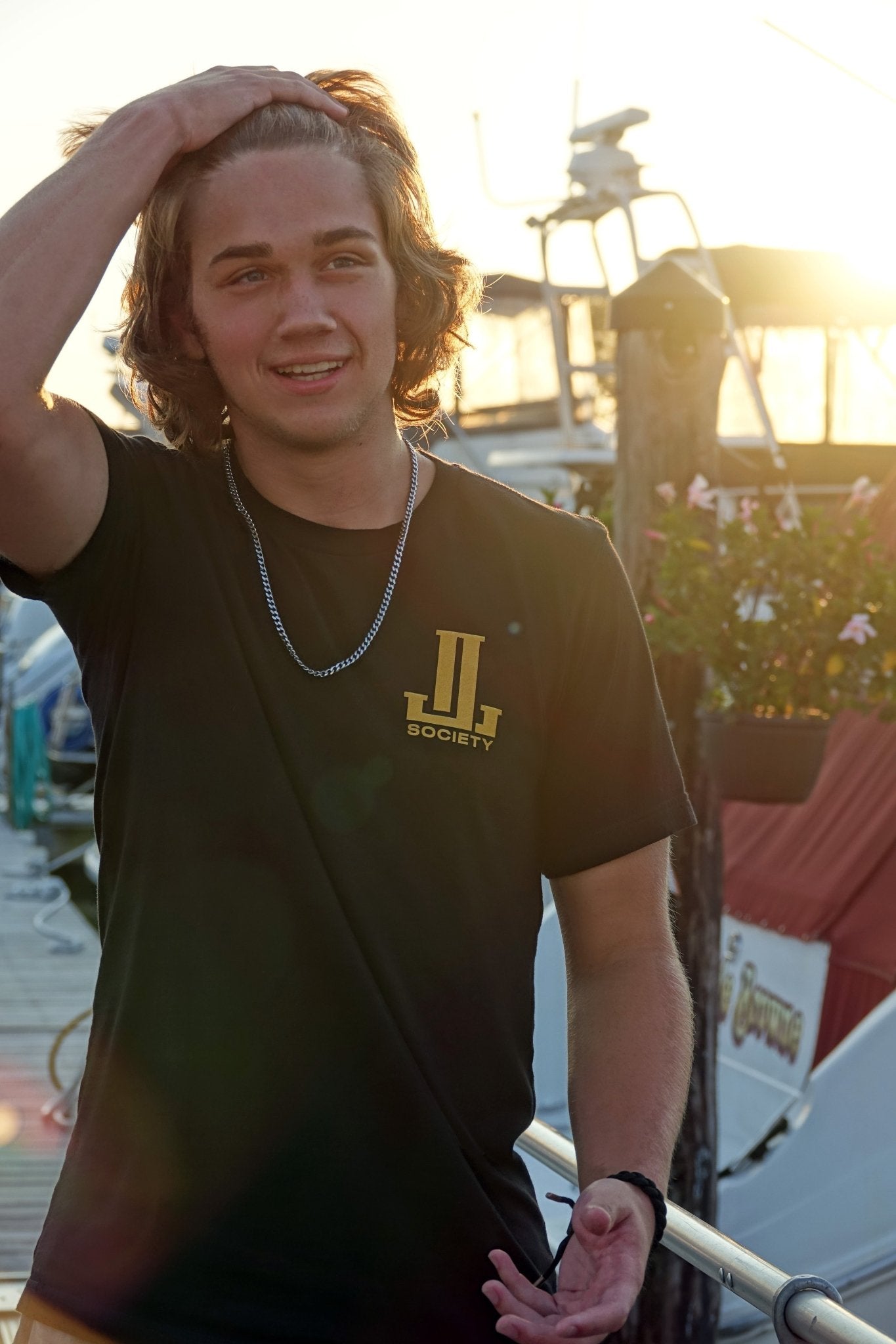 Young man with wavy hair wearing a black t-shirt, smiling at sunset near boats.